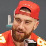 A smiling athlete, Travis Kelce, wearing a red Kansas City Chiefs cap speaks at a press conference. and this blog is exploring a question what is travis kelce net worth