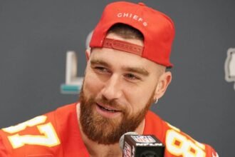 A smiling athlete, Travis Kelce, wearing a red Kansas City Chiefs cap speaks at a press conference. and this blog is exploring a question what is travis kelce net worth